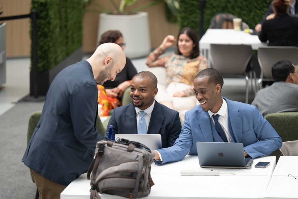 Três homens em trajes sociais conversam e sorriem ao redor de uma mesa com laptops em um ambiente corporativo.