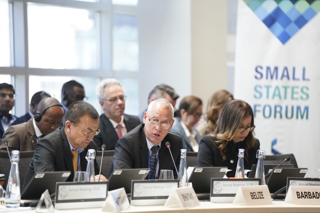 Pessoas em trajes formais participam de uma reunião com a faixa "SMALL STATES FORUM" ao fundo e placas de identificação de países como "BELIZE" e "BARBADOS" sobre a mesa.