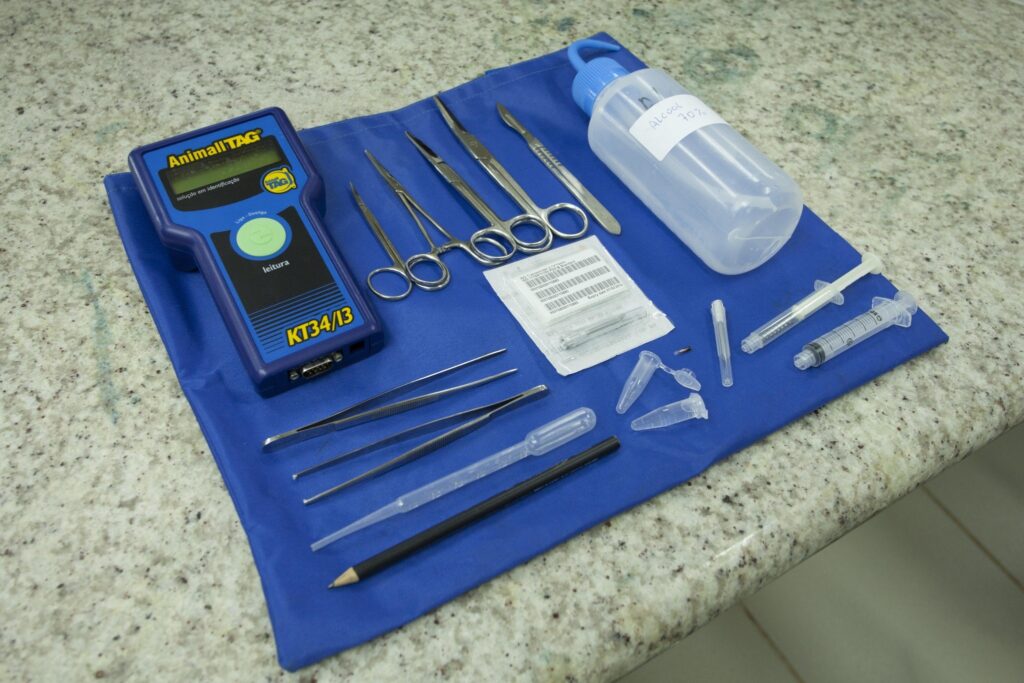 Instrumentos de coleta laboratorial, incluindo leitor 'Animall TAG', seringas, pinças e frasco de 'ALCOOL 70%', sobre um pano azul.