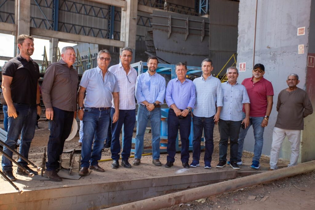 Grupo de dez homens posa em pé em ambiente industrial com estruturas metálicas ao fundo.