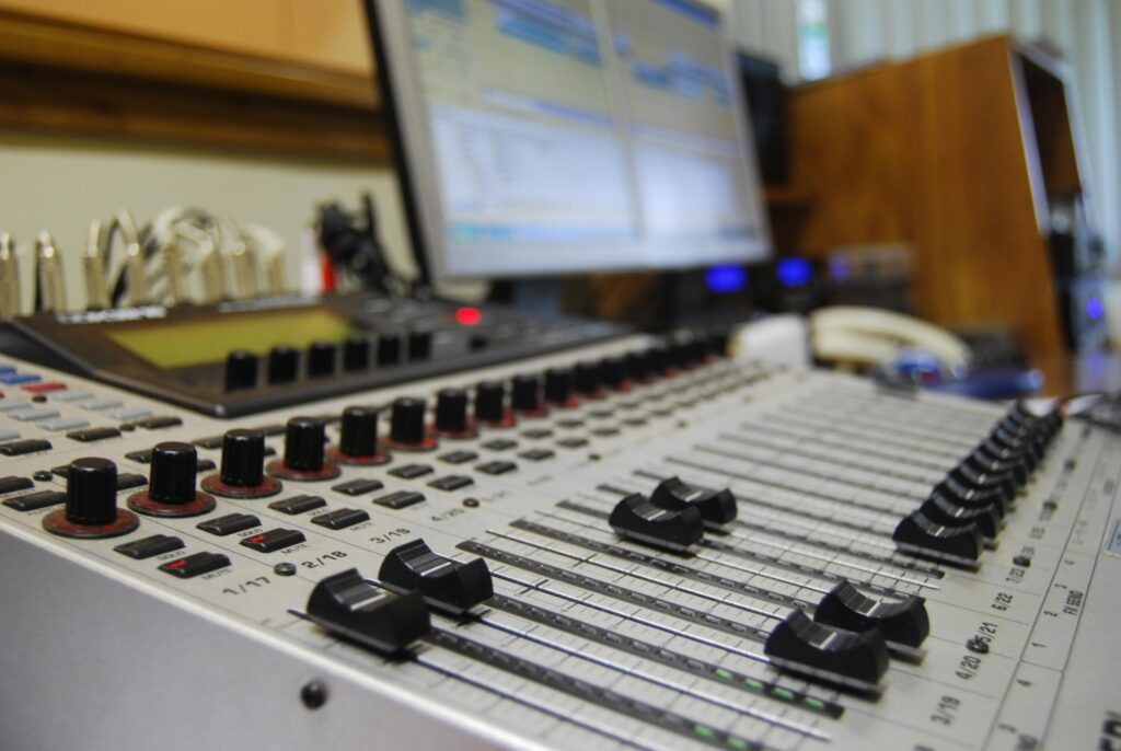 Mesa de mixagem de áudio com controles deslizantes e botões em destaque, com um monitor ao fundo.