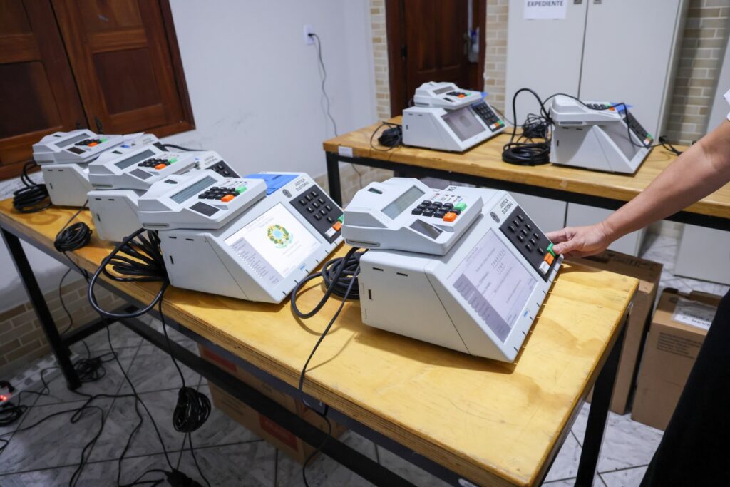 Várias urnas eletrônicas da "Justiça Eleitoral" sobre mesas de madeira, com a mão de uma pessoa tocando o botão verde de um dos aparelhos.