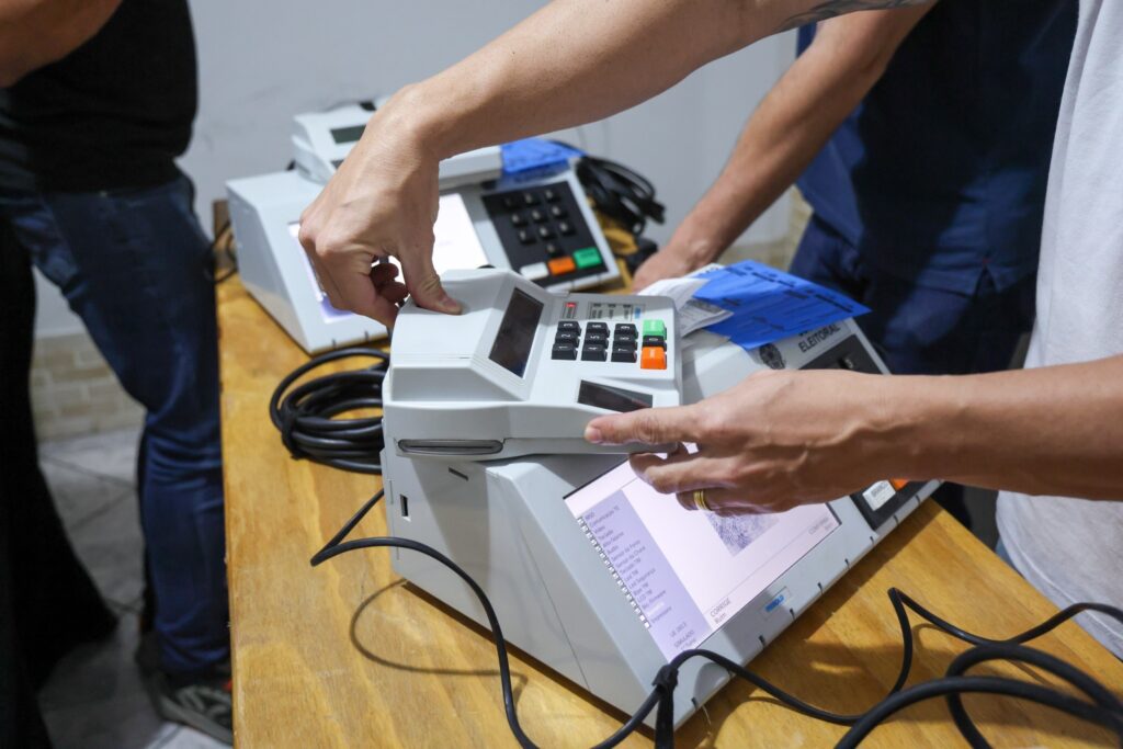 Mãos manuseiam urnas eletrônicas sobre uma mesa de madeira; a tela de uma delas exibe uma lista de verificação técnica e a palavra "SIMULADO".
