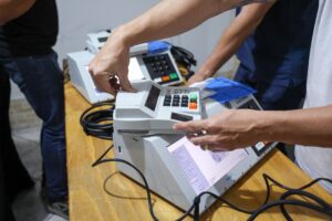 Mãos manuseiam urnas eletrônicas sobre uma mesa de madeira; a tela de uma delas exibe uma lista de verificação técnica e a palavra "SIMULADO".