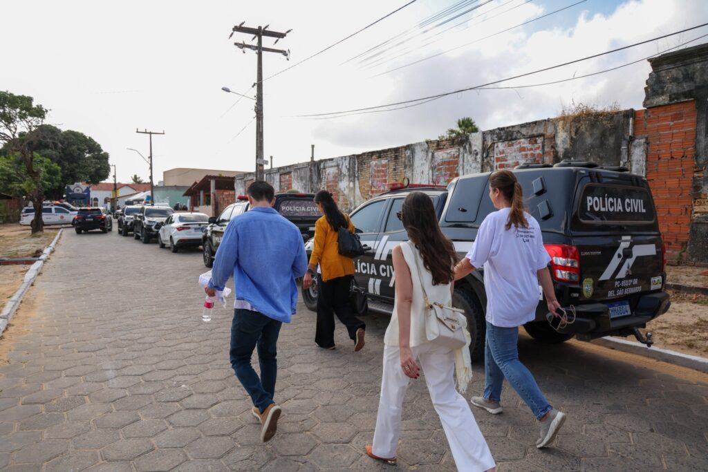 Quatro pessoas caminham por uma rua de pedras ao lado de viaturas da "POLÍCIA CIVIL".