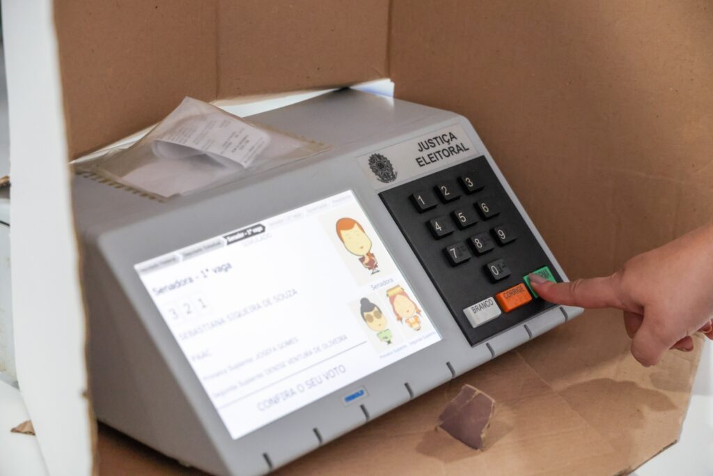 Mão pressiona o botão "Confirma" de uma urna eletrônica que exibe a mensagem "Senadora - 1ª vaga" e o termo "SIMULADO".