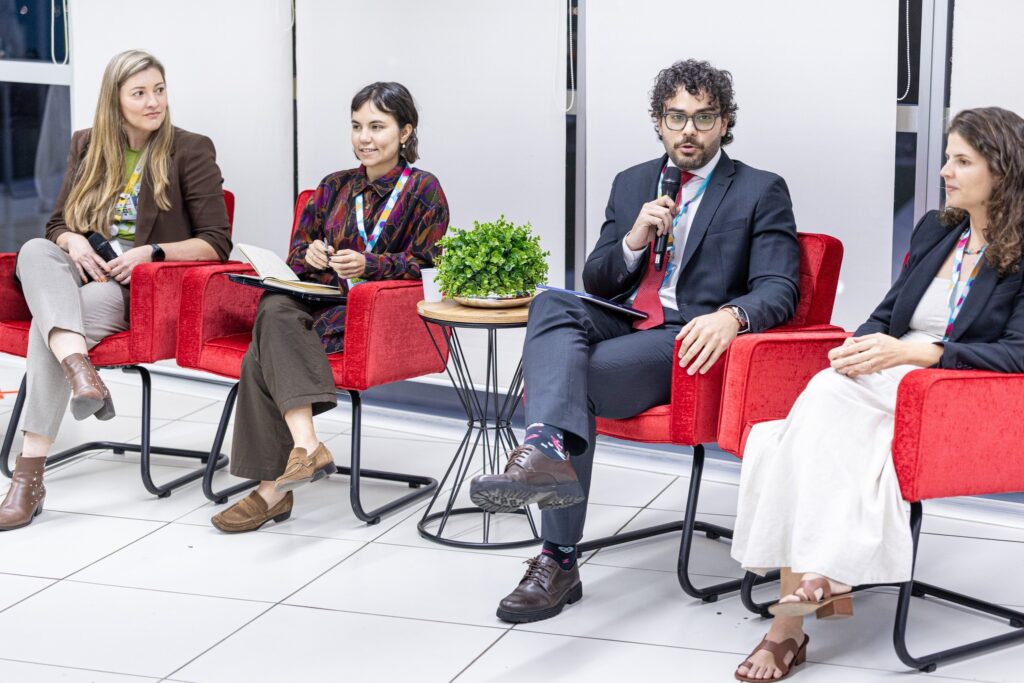 Quatro pessoas participam de painel sentadas em poltronas vermelhas, com um homem ao centro falando ao microfone.