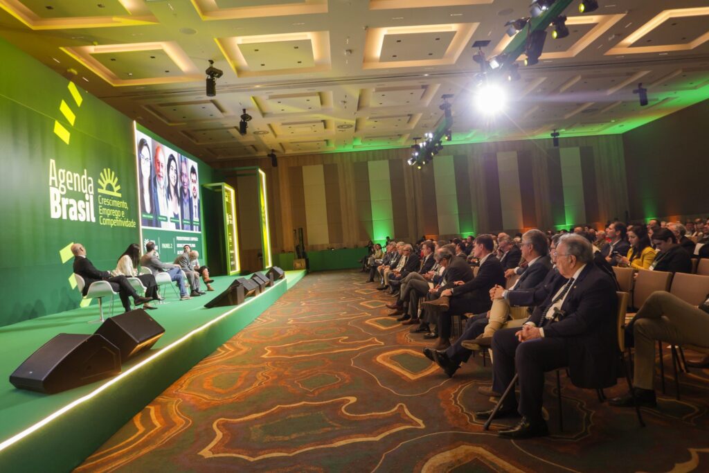 Participantes de um evento sentados em um auditório, com um painel ao fundo com os dizeres "Agenda Brasil Crescimento, Emprego e Competitividade".