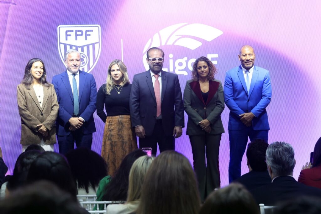 Seis pessoas posam em um palco diante de um painel roxo com os logotipos "FPF" e "Liga F".