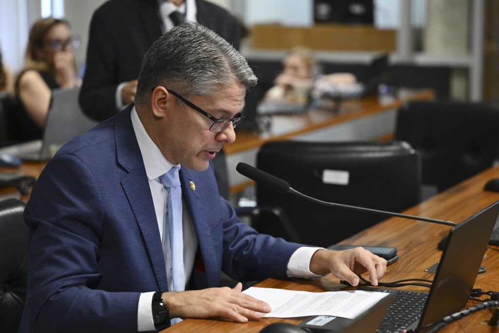 Senador Alessandro Vieira fala ao microfone durante reunião, consultando um documento sobre a mesa.