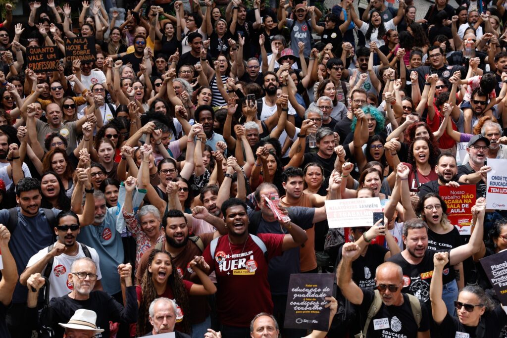 Multidão de professores em manifestação urbana com braços erguidos e cartazes como "Quanto vale um professor?" e "Preencher relatórios também é trabalho".