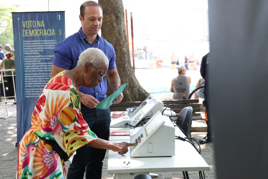 Mulher idosa com vestido floral opera urna eletrônica ao lado de banner com os dizeres "VOTO NA DEMOCRACIA".