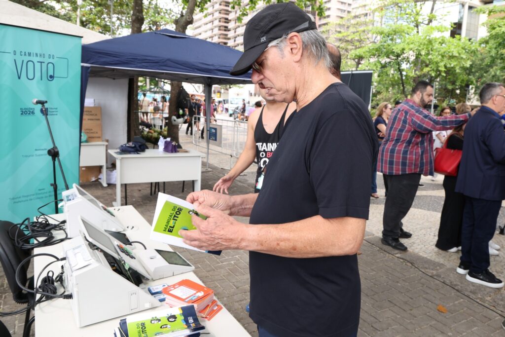 Homem de boné e camiseta preta lê o "MANUAL DO ELEITOR" ao lado de urnas eletrônicas em evento com a faixa "O CAMINHO DO VOTO".