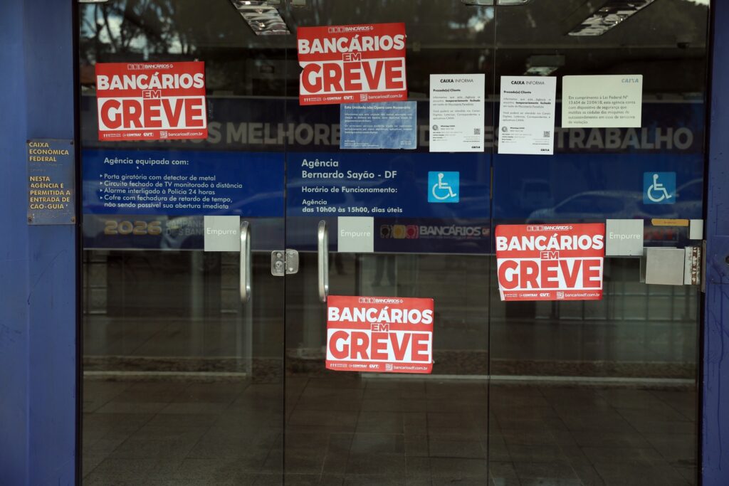Fachada de agência da Caixa Econômica Federal com portas de vidro fechadas e cartazes vermelhos com os dizeres "Bancários em Greve".