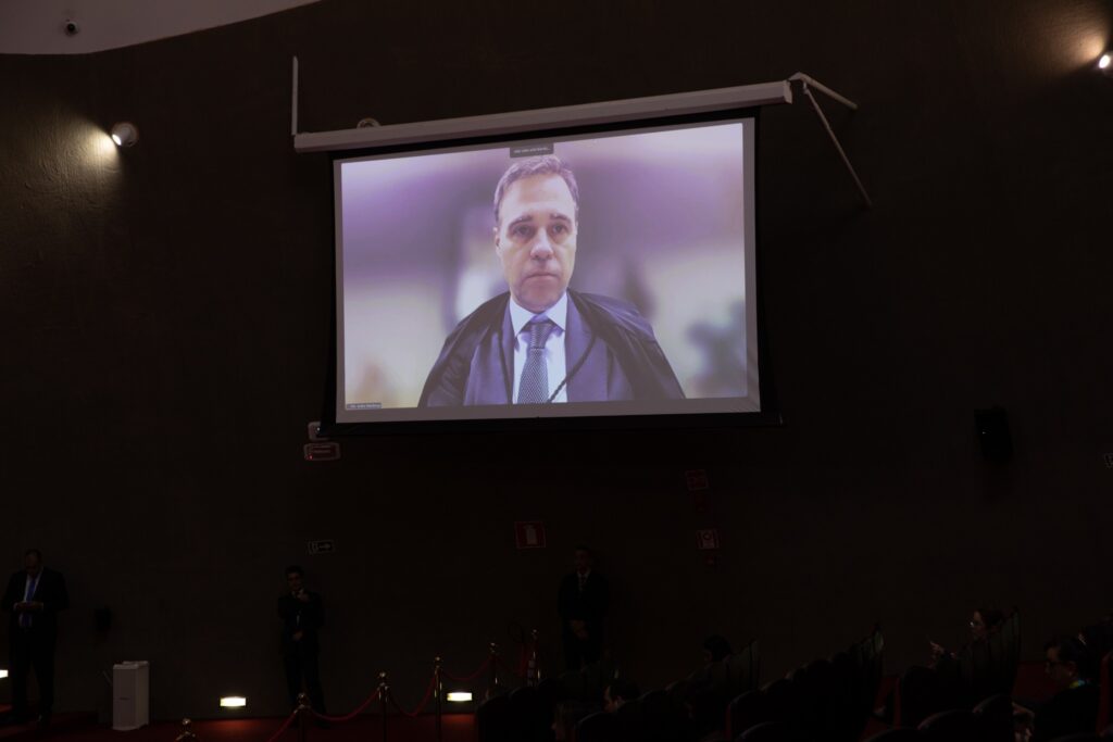 Ministro André Mendonça aparece em uma tela de projeção com a legenda "Min. André Mendonça" durante sessão plenária em ambiente escuro.