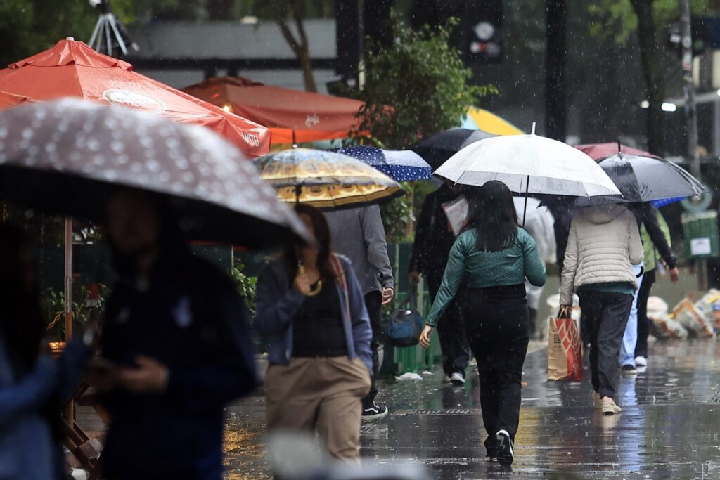 Pedestres caminham sob chuva com guarda-chuvas em calçada urbana molhada em São Paulo.