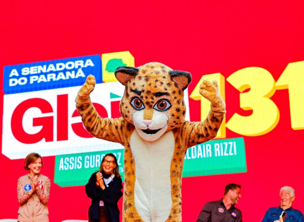 Pessoa fantasiada de onça com braços erguidos em comemoração diante de painel vermelho com inscrições políticas.