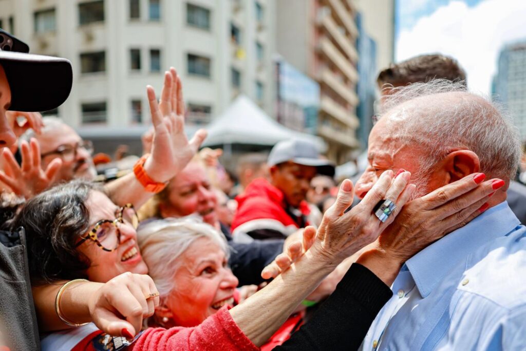 O presidente Luiz Inácio Lula da Silva é abraçado e tem o rosto tocado por apoiadores em ato público em Curitiba.