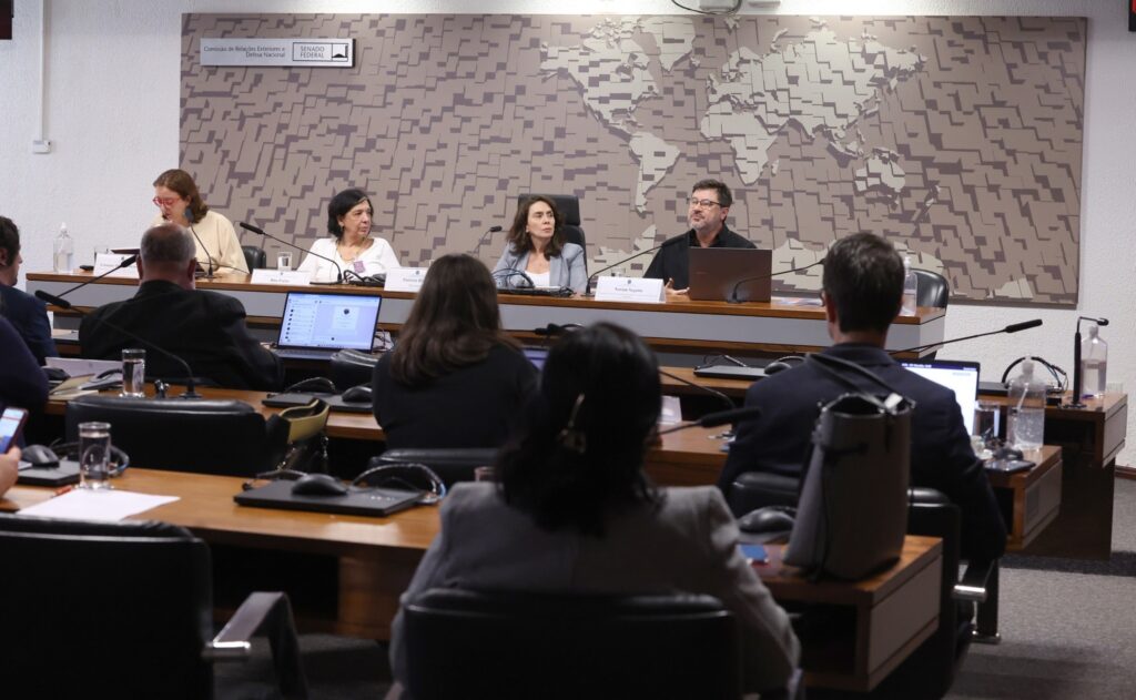 Pessoas em reunião no Senado Federal, com a placa "Comissão de Relações Exteriores e de Defesa Nacional" e um mapa-múndi estilizado ao fundo.