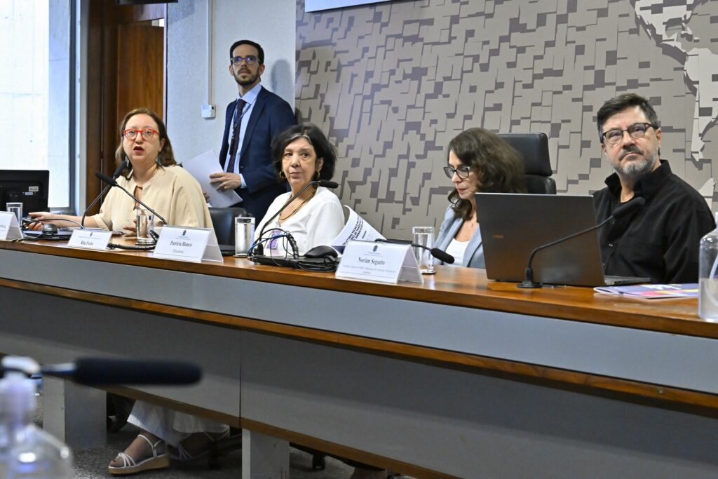 Mesa de reunião com Patrícia Blanco, Rita Freire e Norian Segatto sentados, além de outros participantes.