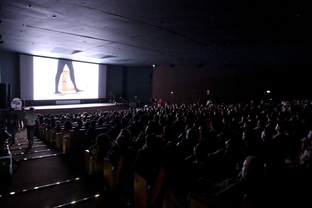 Público lota a sala de cinema escura durante a exibição de um filme na tela grande.