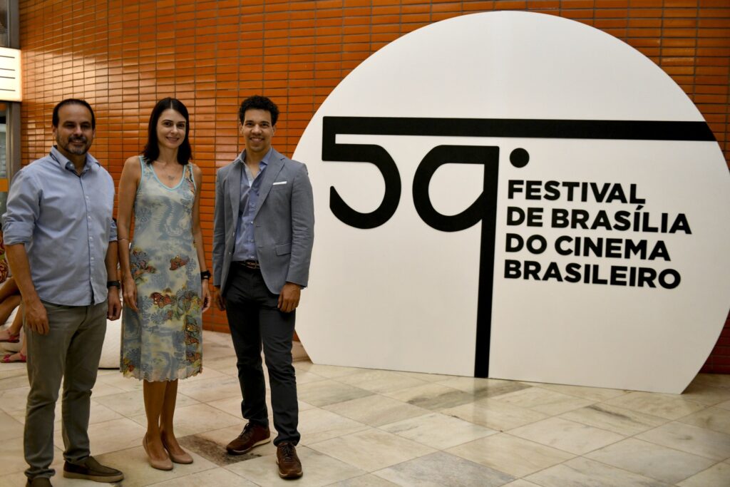 Três pessoas posam sorridentes em pé diante de painel com os dizeres "59º Festival de Brasília do Cinema Brasileiro".