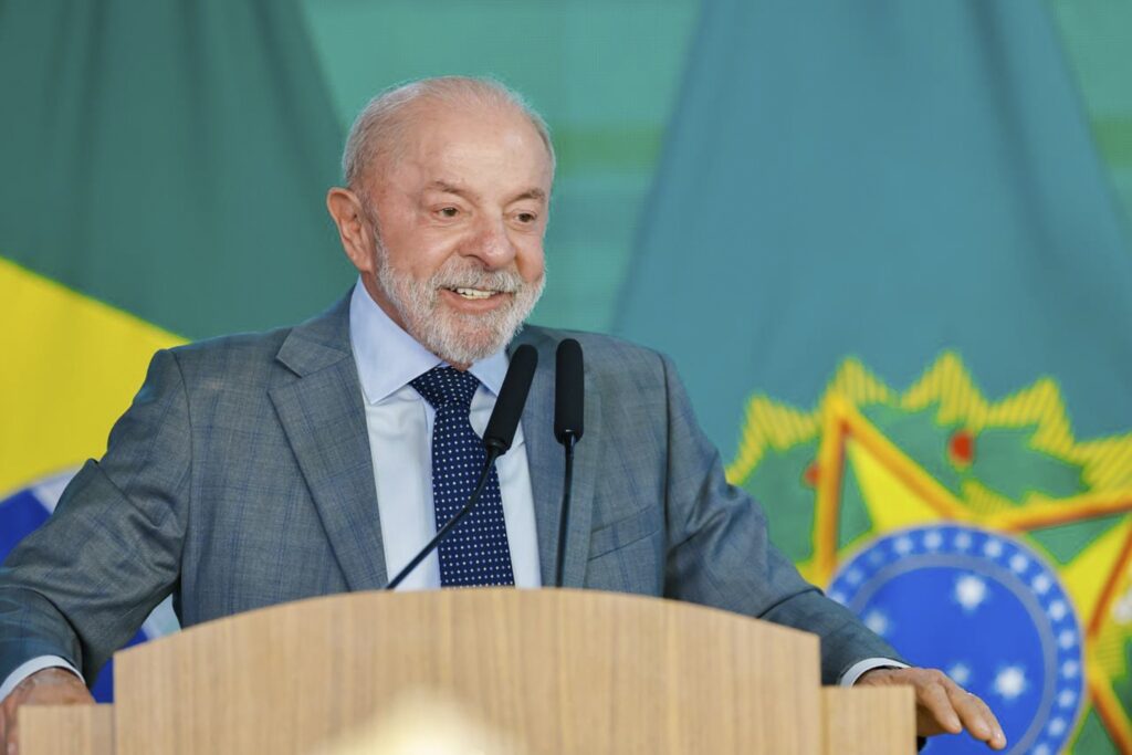 Luiz Inácio Lula da Silva sorri atrás de um púlpito com microfones, com o brasão do Brasil ao fundo.