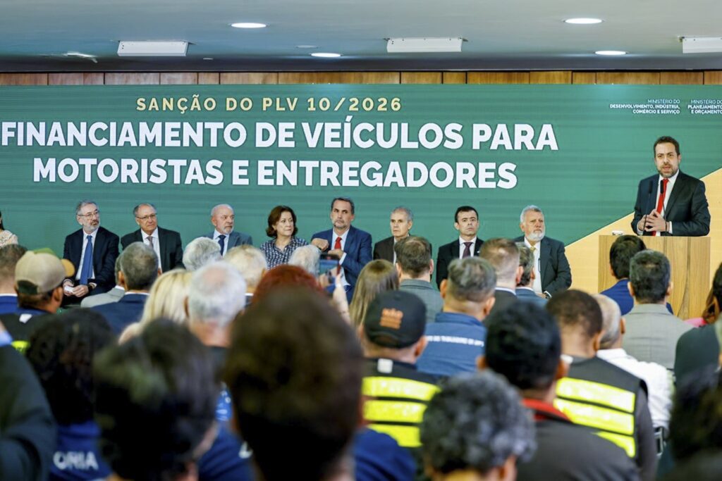 Autoridades sentadas à mesa e homem discursando em púlpito diante de painel com os dizeres "SANÇÃO DO PLV 10/2026 - FINANCIAMENTO DE VEÍCULOS PARA MOTORISTAS E ENTREGADORES".