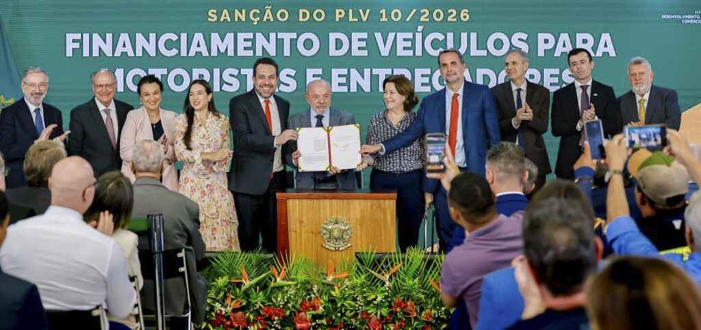 O presidente Luiz Inácio Lula da Silva e autoridades seguram um documento durante a sanção do PLV 10/2026, com o texto "FINANCIAMENTO DE VEÍCULOS PARA MOTORISTAS E ENTREGADORES" ao fundo.