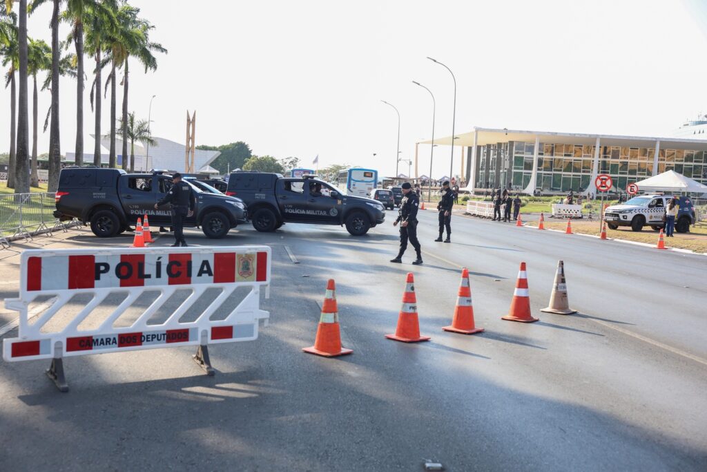 Esquema de segurança com barreiras e viaturas da Polícia Militar próximo ao Congresso Nacional, exibindo placa com os dizeres "POLÍCIA".