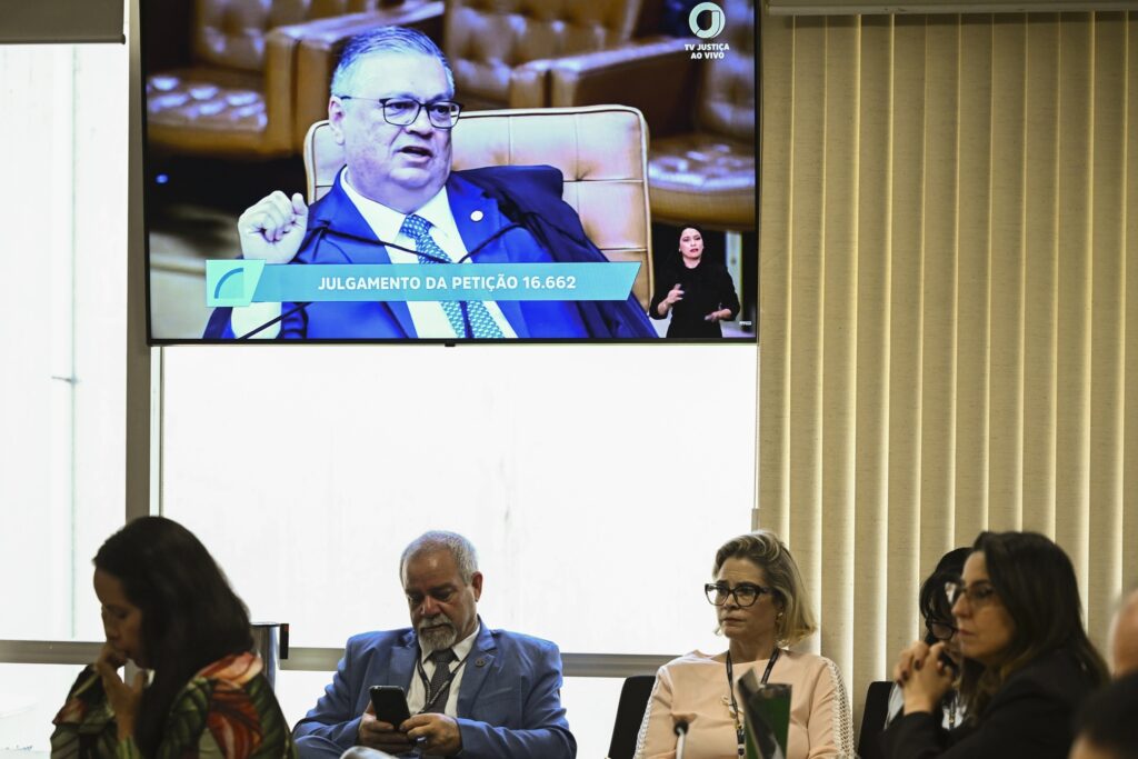 Painel exibe sessão plenária do Supremo Tribunal Federal com julgamento enquanto pessoas acompanham a reunião na mesa.