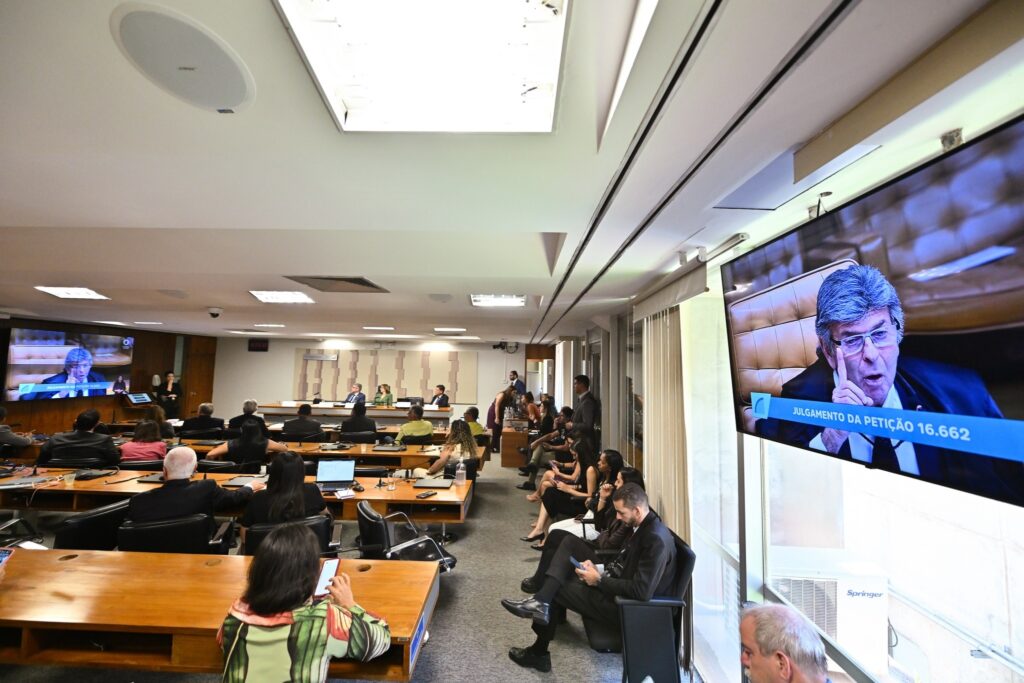 Vista geral de audiência pública em sala legislativa com público sentado às mesas e telão lateral exibindo julgamento no STF.