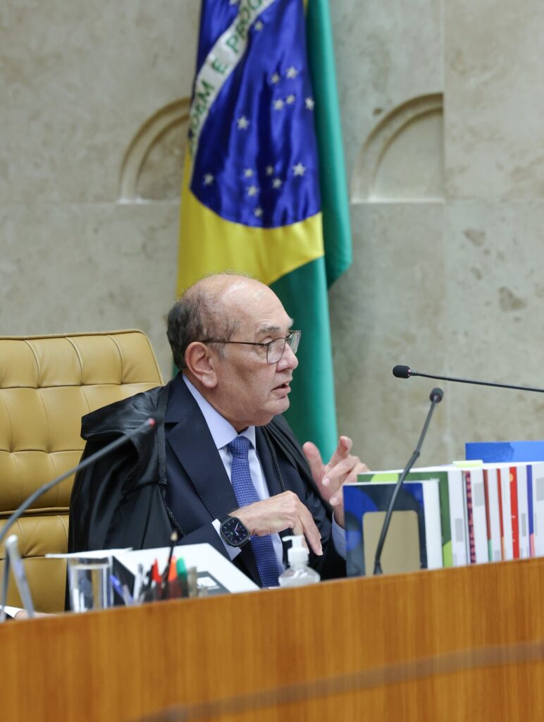 O ministro Gilmar Mendes gesticula durante sessão plenária no Supremo Tribunal Federal, tendo ao fundo a bandeira do Brasil.