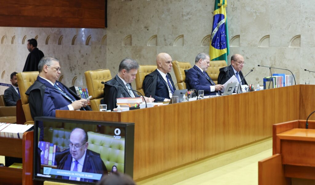 Ministros do STF sentados à bancada durante sessão plenária extraordinária, com a bandeira do Brasil ao fundo.