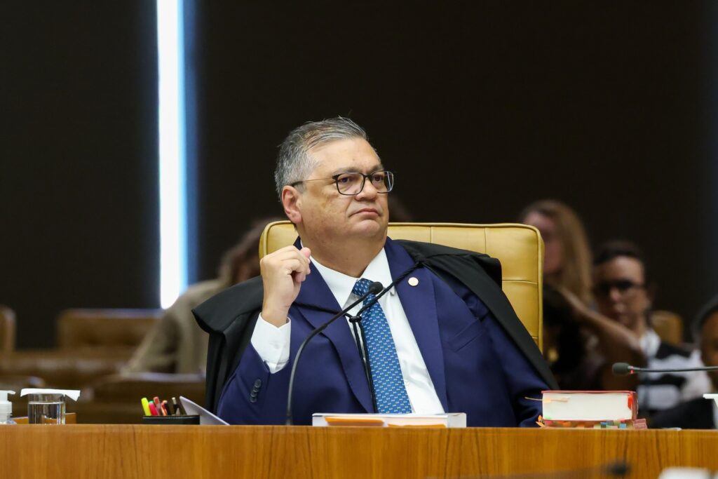 O ministro Flávio Dino sentado à mesa de julgamento no plenário do STF, com expressão séria e usando toga preta.