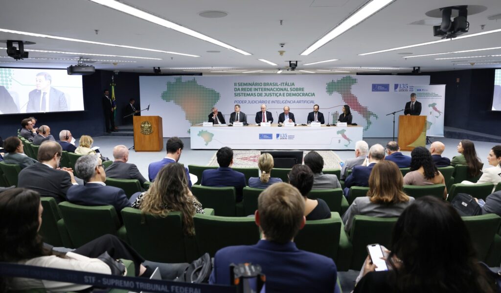 Audiência em auditório assiste ao "II Seminário Brasil-Itália da Rede Internacional Sobre Sistemas de Justiça e Democracia", com autoridades sentadas à mesa e um orador ao pódio.