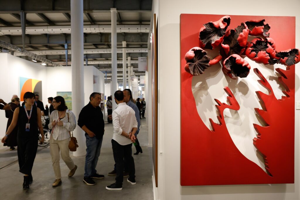 Visitantes em uma galeria de arte observam uma obra tridimensional vermelha e branca na feira ArtRio 2026.