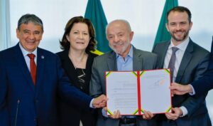 O presidente Luiz Inácio Lula da Silva e outras três autoridades posam sorridentes segurando um documento oficial com capa vermelha, verde e amarela no Palácio do Planalto.