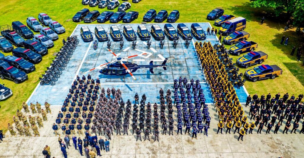 Vista aérea de forças de segurança reunidas em pátio com helicóptero da "PRF" ao centro, cercado por viaturas e policiais em diversas fardas.