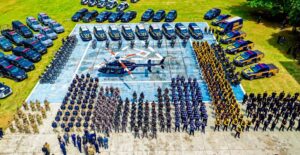 Vista aérea de forças de segurança reunidas em pátio com helicóptero da "PRF" ao centro, cercado por viaturas e policiais em diversas fardas.