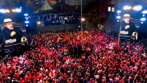Multidão densa de apoiadores com celulares erguidos em comício político noturno, com telas laterais exibindo a imagem de Luiz Inácio Lula da Silva.