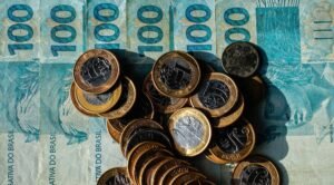comite gestor do ibs: Cédulas de cem reais e moedas brasileiras empilhadas sobre uma superfície.