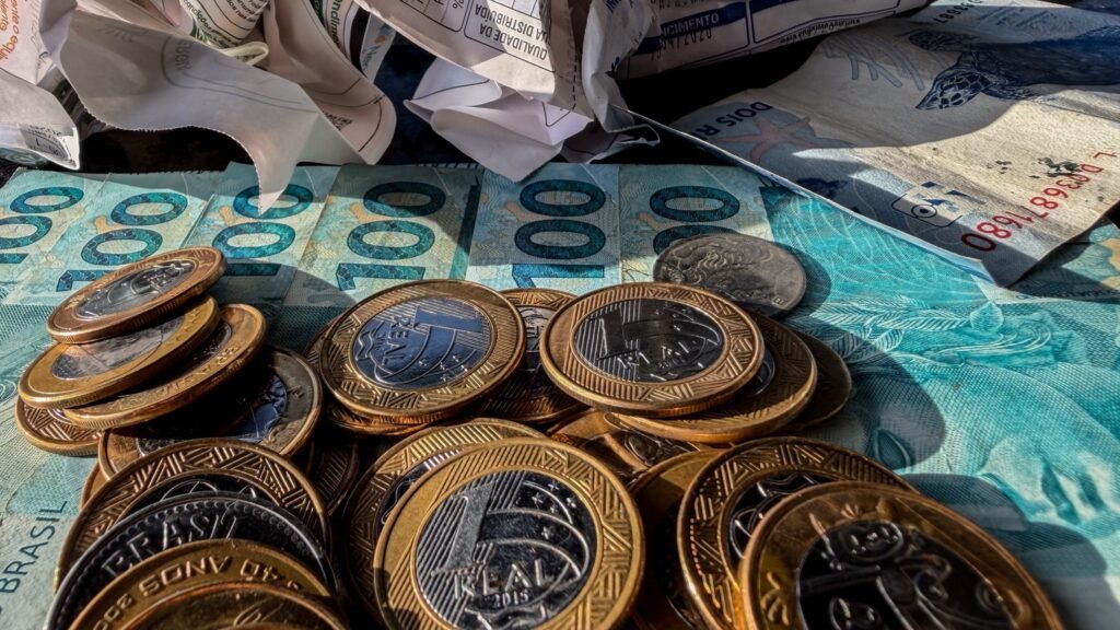 Open Finance: Moedas de 1 real e cédulas de 100 e 2 reais sobre papéis amassados.