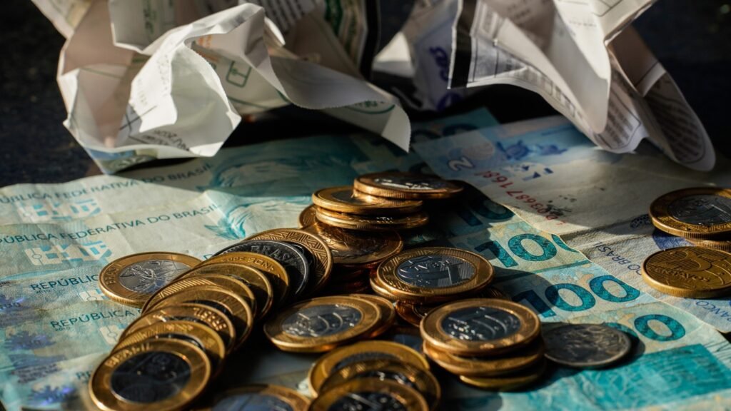 alta do dólar: cédulas e moedas de Real brasileiro espalhadas sobre uma superfície com papéis ao fundo.