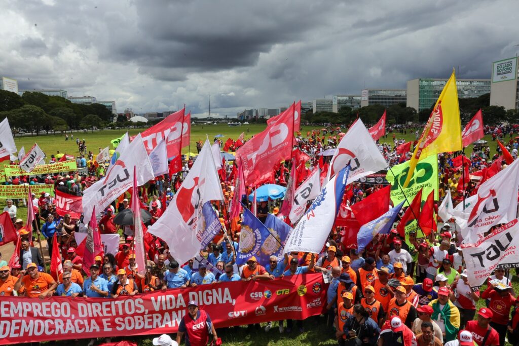 fim da escala 6x1: Manifestantes em área verde seguram diversas bandeiras e uma faixa vermelha com reivindicações da classe trabalhadora.