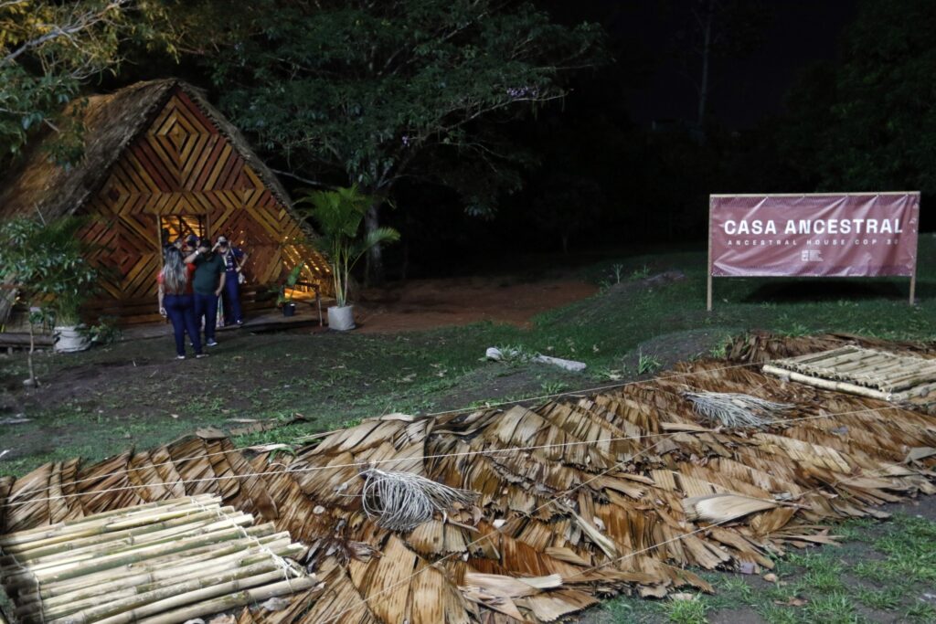saúde indígena no Amazonas: Pessoas reunidas em frente à construção tradicional da Aldeia COP com detalhes em palha e madeira.