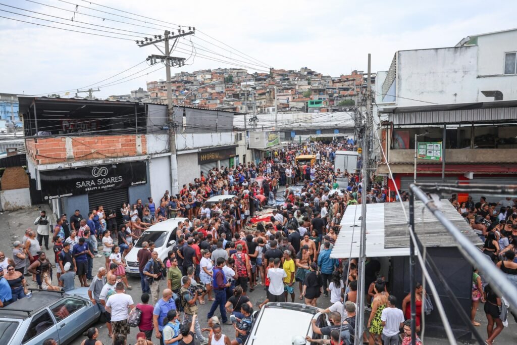 Mãe Bernadete: Multidão reunida em praça urbana ao redor de corpos dispostos no chão, com casas em morro ao fundo.