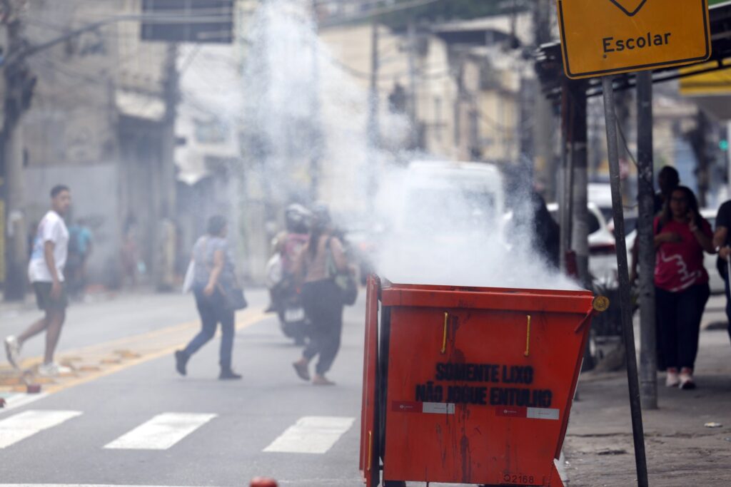 Operação Backbone: lixeira metálica vermelha incendiada exalando fumaça em rua urbana com pedestres desfocados.
