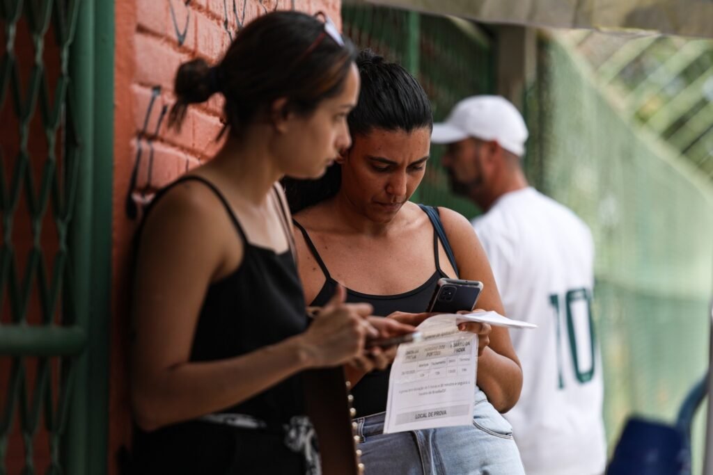 Prova Nacional Docente: duas mulheres consultam documento de local de prova e smartphone em área externa.