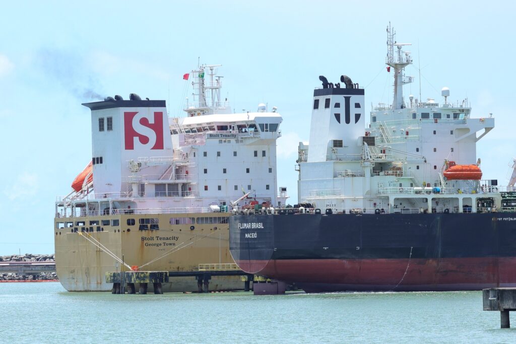 Dois grandes navios cargueiros, o "Stolt Tenacity" e o "Flumar Brasil", atracados em um porto sob céu azul.